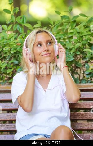 Die entspannte Frau schaut weg und lächelt, während sie Musik hört, mit Kopfhörern, die auf einer Bank in einem Park sitzen. Technik und Freizeitkonzept. Stockfoto