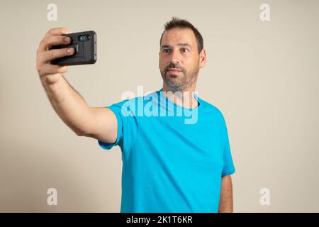 Porträt eines bärtigen spanischen Mannes, der ein Selfie mit seinem Smartphone auf beigefarbenem Studiohintergrund gemacht hat. Stockfoto