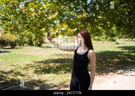 Fröhliche junge Frau in legerer Kleidung, die lächelt und Selfie per Smartphone nimmt, während sie am Sommerwochenende auf der sonnenbeleuchteten Parkallee steht Stockfoto
