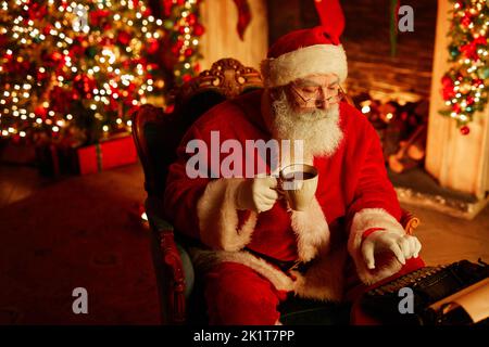 Hochwinkelporträt des traditionellen Weihnachtsmannes mit Schreibmaschine und genießen heiße Schoko am Weihnachtsabend, kopieren Raum Stockfoto