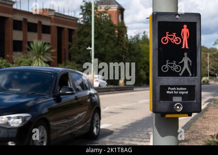 Crosswalk-Taste für Fußgänger und Radfahrer mit Lichtwarnung in Großbritannien. Die Anzeige leuchtet rot. Ein Auto fährt vorbei, verschwommener Hintergrund. Stockfoto