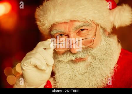 Nahaufnahme eines lächelnden Weihnachtsmanns, der zu Weihnachten auf die Kamera schaut und die Brille justiert Stockfoto