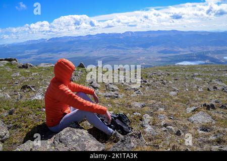 Eine Frau in einer orangefarbenen Jacke mit Kapuze sitzt für eine Pause mit Blick auf die Berge nahe der Spitze des Mount Elbert, dem höchsten Gipfel in Colorado USA Stockfoto