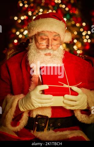 Vertikales Porträt von bärtigen traditionellen Weihnachtsmann Eröffnung Geschenk mit Licht im Inneren von Weihnachtsbaum Stockfoto
