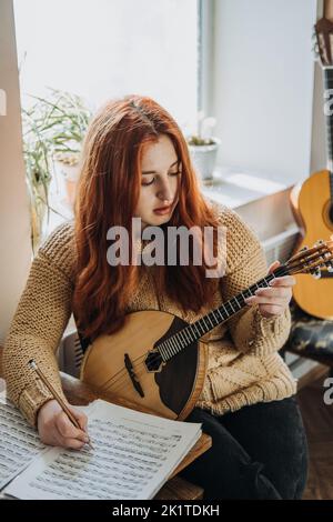 Redhead Frau spielt akustisches Domra Folk Musical Instrument und schaut in die Note. Junge Frau, die zu Hause Musikunterricht nimmt Stockfoto