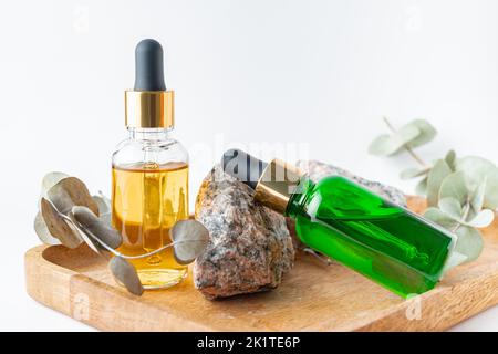 Tropfflaschen aus Glas mit einer Pipette mit goldener Gummispitze und grünen Eukalyptusblättern daneben auf einem Holztablett auf weißem Grund. Naturhautkonzept Stockfoto