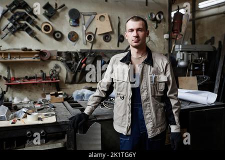 Porträt eines jungen männlichen Arbeiters, der in der Werkstatt steht und Kamera, Werkzeuge im Hintergrund und Kopierraum ansieht Stockfoto