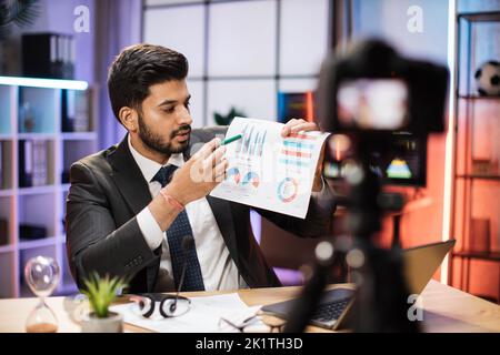 Erfolgreicher arabischer Geschäftsmann oder Makler in formellem Anzug sitzt vor der Kamera auf einem Stativ im Abendbüro und erklärt seinem Publikum Grafiken und Diagramme mit Diagramm. Stockfoto