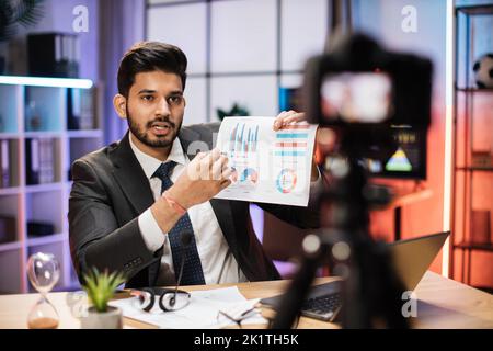 Erfolgreicher arabischer Geschäftsmann oder Makler in formellem Anzug sitzt vor der Kamera auf einem Stativ im Abendbüro und erklärt seinem Publikum Grafiken und Diagramme mit Diagramm. Stockfoto