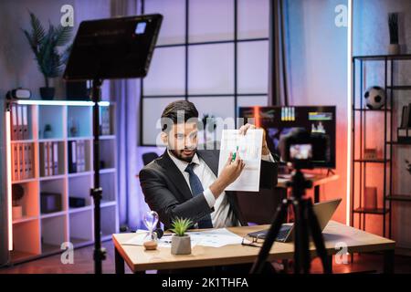 Selbstbewusster, qualifizierter arabischer Händler oder Büroangestellter, der bei Online-Meetings vor der Videokamera sitzt und den Bericht mit Grafiken und Diagrammen erklärt. Stockfoto