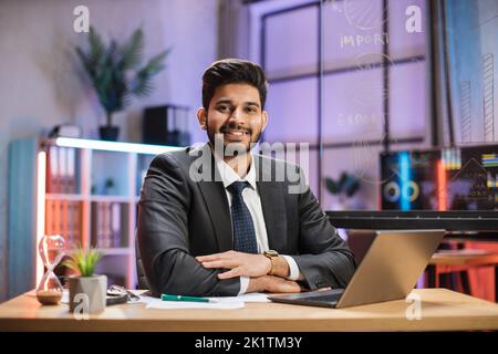 Vorderansicht Porträt von jungen lächelnden erfahrenen smart stilvollen bärtigen hinduistischen Büromanager in Anzug sitzen am Tisch mit Laptop in Abendbüro. Stockfoto
