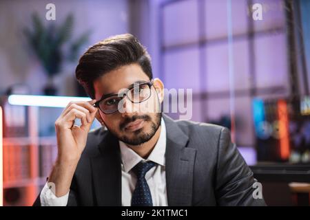 Nahaufnahme eines selbstbewussten indischen Geschäftsmanns oder Wirtschaftswissenschaftlers in formeller Kleidung und Brille, der im Nachtbüro vor der Kamera posiert. Stockfoto