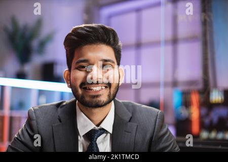 Lächelnder, positiver, selbstbewusster indischer Mann in formeller Kleidung, der am Schreibtisch sitzt. Firmenmitarbeiter mit Bart und Blick auf die Kamera. Konzept von Menschen und Unternehmen. Stockfoto