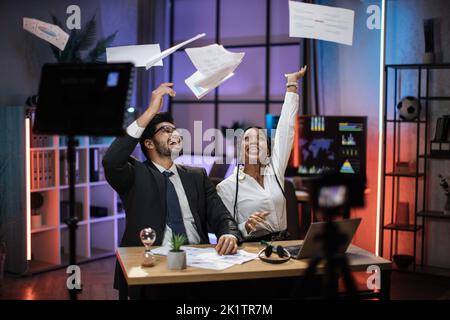 Überglücklich multirassische Geschäftsleute, afroamerikanische Geschäftsfrau und indischer Makler, die im Abendbüro gute Nachrichten erhalten und Zeitungen in die Luft werfen, feiern und nach der Arbeit Spaß haben. Stockfoto
