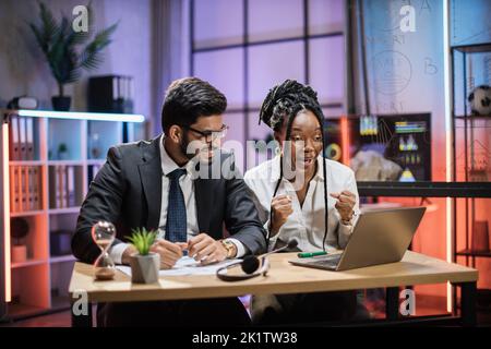 Überglücklich multirassische Geschäftsleute, afroamerikanische Geschäftsfrau und indischer Broker, die im Abendbüro gute Nachrichten erhalten und auf den Laptop-Bildschirm schauen. Stockfoto