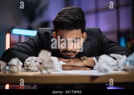 Porträt eines müden, überarbeiteten bärtigen arabischen Geschäftsmannes in formellem Anzug, der im Abendbüro mit zerknitterten Papierblättern auf dem Schreibtisch schlief. Stockfoto