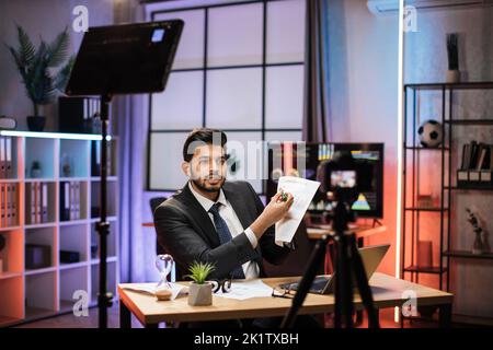 Erfolgreicher arabischer Geschäftsmann oder Makler in formellem Anzug sitzt vor der Kamera auf einem Stativ im Abendbüro und erklärt seinem Publikum Grafiken und Diagramme mit Diagramm. Stockfoto