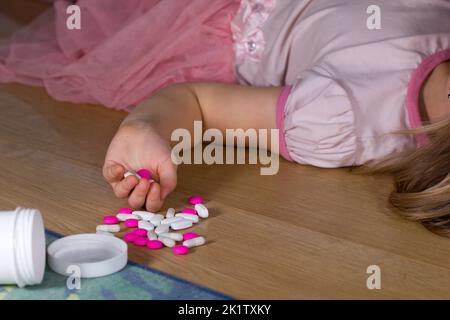 Unbewusstes kleines Kind (Mädchen) mit rosa Pillen, die zu Hause auf dem Boden liegen. Gefahr der Medikamentenvergiftung. Stockfoto