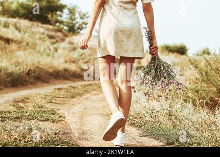 Ein Mädchen mit einem Blumenstrauß aus Herbstblumen geht mit dem Rücken zur Kamera auf einer herbstlichen Straße entlang. Herbstlandschaft Stockfoto