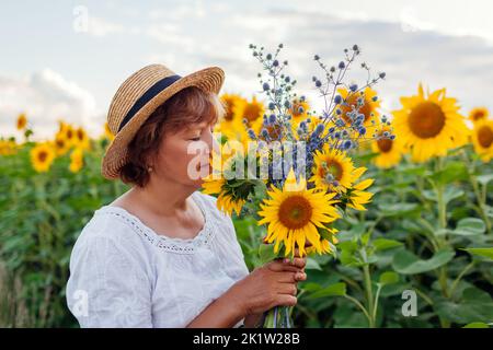 Ältere Frau im Strohhut riecht im Sommerfeld bei Sonnenuntergang nach gelben Sonnenblumen. Frau mittleren Alters bewundert Blüten, die sich im Freien entspannen Stockfoto