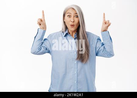 Überraschte alte Frau, asiatische Dame 50 Jahre alt, keuchend erstaunt, zeigt die Finger nach oben, zeigt Werbung, stehend über weißem Studio-Hintergrund Stockfoto