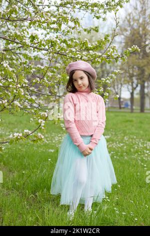 Niedliches kleines Mädchen steht auf dem grünen Gras neben einem blühenden weißen Apfelbaum im Park. Stockfoto