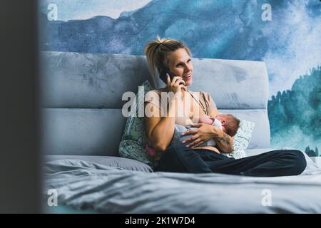 Weiße Frau mit blonden Haaren sitzt im Bett und stillt ihr neugeborenes Baby, während sie am Telefon spricht. Beschäftigt Mama. Verantwortung. Horizontale Innenaufnahme. Hochwertige Fotos Stockfoto