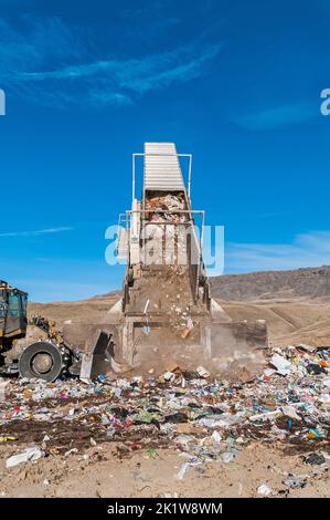 Ein Anhänger von einem Sattelschlepper wird in einem Müllkipper hochgehoben und wirft Müll aus, während eine Bodenverdichter-Planierraupe den Müll schiebt. Stockfoto