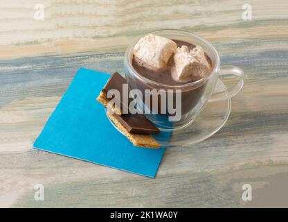 Heiße Schokolade in durchsichtiger Tasse mit schmelzenden Marshallows und Schokolade auf graham Crackers Snack auf Untertasse auf blauer Serviette Stockfoto