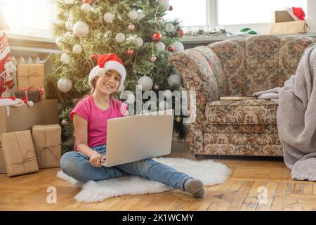 Ein glückliches kleines Mädchen öffnet zu Hause unter dem Weihnachtsbaum ein Geschenk und kommuniziert mit ihren Großeltern über einen Laptop via Videolink. Urlaubskonzept Stockfoto