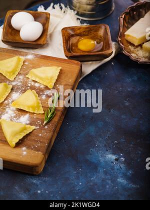 Kochen hausgemachte italienische Ravioli. Rohe Ravioli auf einem Holzschneidebrett auf blauem Marmorhintergrund. Es ist freier Speicherplatz zum Einfügen vorhanden. Rezepte für Restau Stockfoto