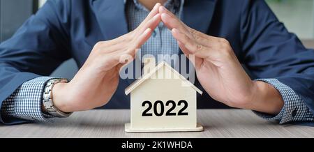 Geschäftsmann übergibt Holzhaus Modell mit 2022 Neujahr Text. Immobilienversicherung und Immobilienkonzepte Stockfoto