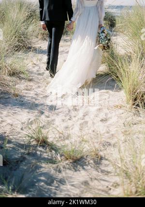 Ein Bräutigam und eine Braut, die an einem Sandstrand mit wildem Gras, vertikaler Aufnahme, spazieren gehen Stockfoto