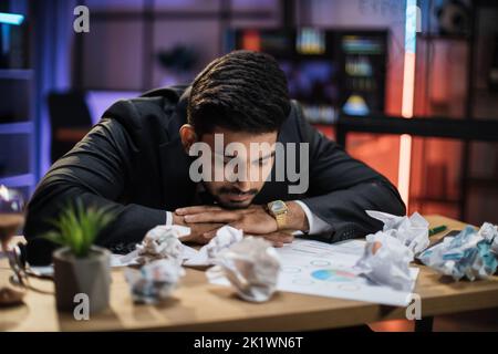 Porträt eines müden, überarbeiteten bärtigen arabischen Geschäftsmannes in formellem Anzug, der im Abendbüro mit zerknitterten Papierblättern auf dem Schreibtisch schlief. Stockfoto