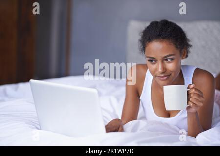 Auf dem Laufenden bleiben mit lokalen Nachrichten. Eine junge Frau genießt eine Tasse Kaffee, während sie an einem Laptop arbeitet. Stockfoto