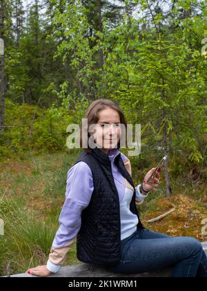 Wanderer, der während eines Waldspaziergangs Pause einnahm. Junge kaukasische Frau, die in einem grünen Wald auf einem umgestürzten Baum sitzt und das Telefon in der Hand hält. Technologiekonzept Stockfoto