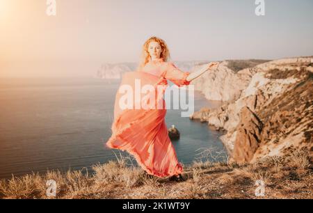 Lockig rothaarige kaukasische Frau mit Sommersprossen. Süße schöne junge blonde Frau in einem rosa langen Kleid posiert auf einem vulkanischen Felsen hoch über dem Meer Stockfoto