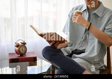 Cropped Shot, Ein lässig-hübscher junger asiatischer Mann, der ein Buch liest, während er im Café einen Kaffee schlürft. Stockfoto