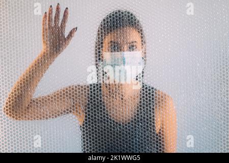 Selbstisolierung. Unschärfes Hochformat. Einsamkeit unter Quarantäne stellen. Gestörte Frau in schützende Gesichtsmaske hinter transparenten Polyethylen Luftpolsterfolie gefangen Stockfoto