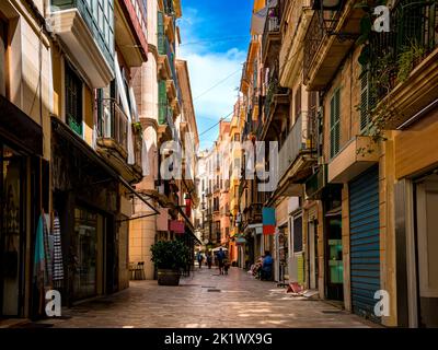 Wenige Touristen laufen durch eine enge mediterrane Gasse mit kleinen Geschäften und Restaurants im Schatten von Wohngebäuden im Zentrum von Palma. Stockfoto