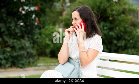 Kaukasische Brünette Frau in weißem Kleid sitzt auf der Bank im Stadtpark. Mädchen, die begeistert auf dem Handy reden. Seitenansicht Wetter Bewölkt Sommertag Stockfoto