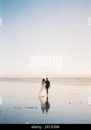 Eine schöne vertikale Rückaufnahme eines frisch verliebten Paares, das barfuß am Strand läuft Stockfoto