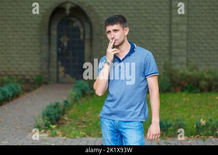 Ein unrasierter Mann steht auf der Straße und raucht in Erwartung von Gedanken und Erfahrungen, tagsüber in einem blauen T-Shirt und Jeans vor dem Hintergrund eines Steinzauns Stockfoto