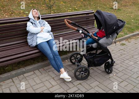 Eine junge Frau schläft auf einer Bank neben dem Kinderwagen, während sie mit ihrem kleinen Kind geht Stockfoto