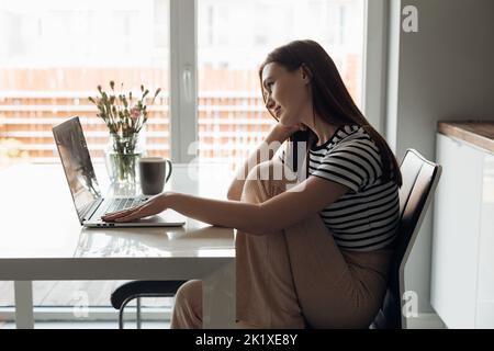 Gelangweilte, ruhige junge, braune Frau, die mit gebeugten Beinen in der Küche sitzt, am Laptop arbeitet und Kaffee in der Nähe des Fensters trinkt Stockfoto