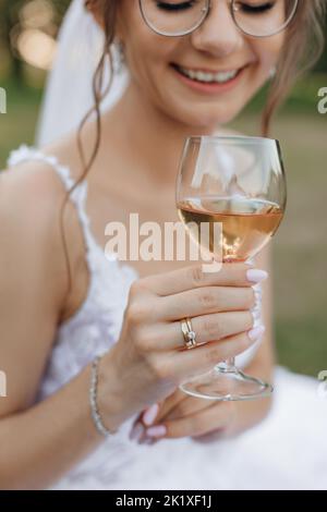 Vertikales Porträt einer zugeschnittenen lächelnden Braut im Hochzeitskleid und Schleier mit Gläsern, die Sekt in Weinglas trinken Stockfoto