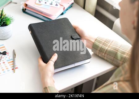 Nahaufnahme eines Frauenhands mit schwarzem Mockup-Notizbuch. Mädchen hält einen schwarzen leeren Notizblock mit einem kleinen Kaktus auf dem Tisch. Mockup leer Stockfoto
