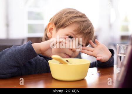 Kleiner Junge sitzt am Tisch und mit Interesse beobachten Cartoon auf Tablet-Computer Nahaufnahme. Kinder im Kindergartenalter essen Schokoladenkugeln mit Milch. Autismus Stockfoto