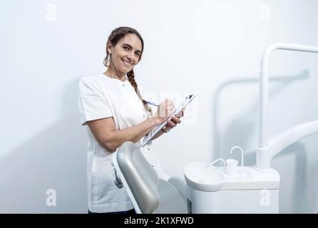 Zahnarzthelferin oder Zahnärztin auf dem Hintergrund des Zahnarztzimmers. Porträt einer jungen Ärztin in weißer Uniform. Hübsche Krankenschwester ist lo Stockfoto