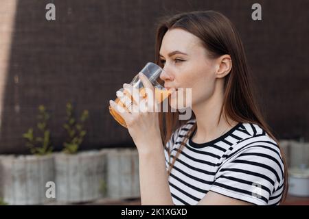 Junge Brünette, ruhige, entspannte Brünette Frau im gestreiften T-Shirt trinken frischen Bio-Orangensaft im Glas draußen Stockfoto